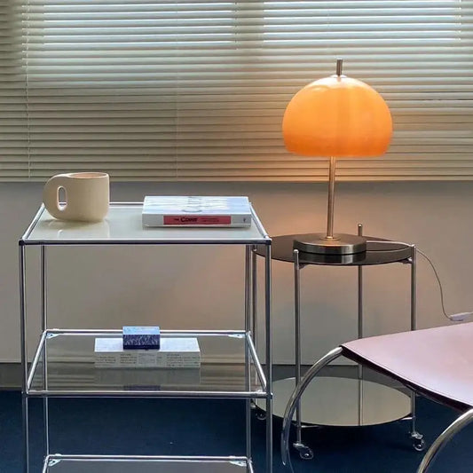 Modern glass side table with a lamp and books against a neutral wall.