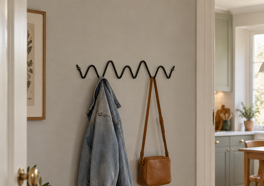 A black metal zigzag wall hook rack mounted on a light gray wall in a home entryway, holding a light blue denim jacket and a brown leather shoulder bag. Below the rack is a woven seat bench with a plaid throw blanket and a pair of beige slippers on the floor.